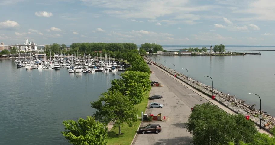 Aerial view of a marina on a large lake with dozens of sailboats and motorboats docked in slips. A tree-lined parking area runs along the marina, while a long rock-lined pier with light poles extends into the water toward a small lighthouse at the harbor entrance under a partly cloudy sky.